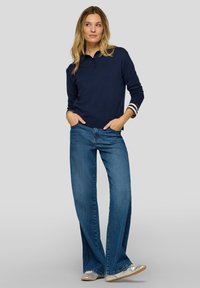 Marineblauer Pullover mit Kragen und drei Knöpfen, kombiniert mit hellblauen Schlagjeans. Der Pullover hat gestreifte Akzente an den Ärmeln.