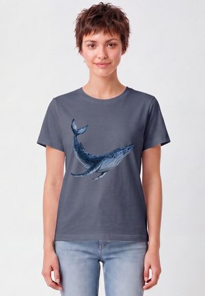 Jong persoon met kort haar draagt een grijs T-shirt met een gedetailleerde illustratie van een blauwe vinvis, gecombineerd met lichtblauwe jeans.