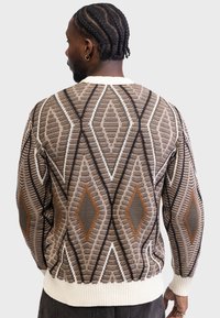 Knit sweater featuring a geometric diamond pattern in brown, beige, and black with a ribbed white hem and cuffs. Unisex design with textured fabric.