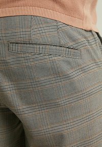 Detailní záběr na šedé a hnědé kostkované kalhoty s zadními kapsami, které jsou kombinovány s broskvově oranžovým žebrovaným svetr.