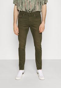Jeans ajustados en verde oliva con diseño de cinco bolsillos y detalles de costura en rojo contrastante, combinados con zapatillas blancas.