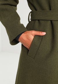 Olivgrön kappa i texturerat tyg med bältesmidja och sidoficka. Ärmens manschett har en svart stickad kant för extra detalj.