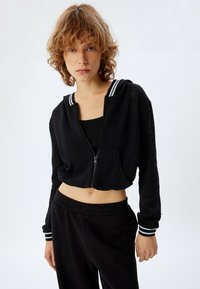 Felpa cropped nera con zip e accenti a righe bianche, realizzata in materiale a maglia testurizzata, abbinata a pantaloni neri con coulisse.