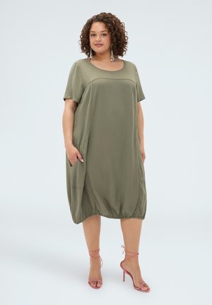 Frau mit lockigem Haar, die ein olivgrünes, lockeres Kleid mit Taschen und pinken High-Heel-Sandalen trägt und vor einem einfarbigen Hintergrund steht.