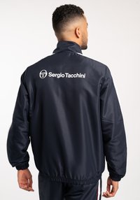 Giacca leggera color blu navy con colletto alto e polsini elasticizzati. Presenta una scritta del logo bianca sulla parte posteriore e strisce laterali a contrasto.