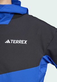 Blauwe en zwarte outdoorjas met een opvallend geborduurd "TERREX"-logo, contrasterende kleuren en een gestructureerde materiaalfinish.