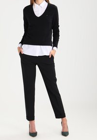 Schwarzer V-Ausschnitt-Pullover über einem weißen Hemd, kombiniert mit schwarzen, schmal zulaufenden Hosen und grünen High Heels, was einen eleganten monochromen Look schafft.