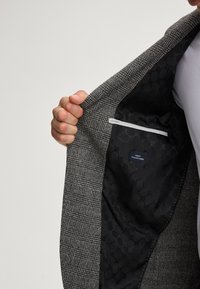 Grijze blokjesjacket met een gladde zwarte voering die een subtiel patroon vertoont. Interne zak met een label dat "Finest Italian Fabric" zegt.