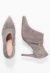 Grå mocka högklackade ankelstövlar med spetsiga tår, rynkdetalj, en sidodragkedja och en guldig hjärtformad accent. Slät yta.