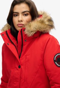 Parka roja con capucha de piel sintética, cierre de cremallera, acentos negros y parche con el logotipo en la manga. Cuenta con bolsillos con cremallera y de solapa.