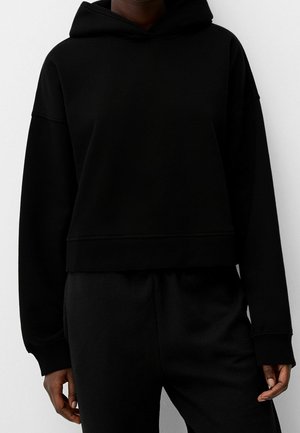 Personne portant un sweat à capuche court noir uni à manches longues et un pantalon noir assorti, debout sur un fond blanc.