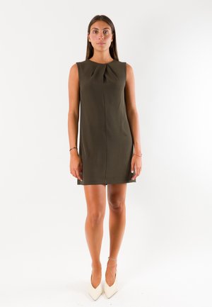Donna in piedi, rivolta in avanti, che indossa un vestito verde oliva senza maniche e tacchi alti bianchi a punta su sfondo bianco.