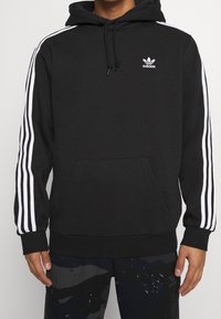 Schwarzer Kapuzenpullover mit weißem Drei-Streifen-Design an den Ärmeln, verstellbarer Kapuze und vorderer Kängurutasche. Mit kleinem Adidas-Logo auf der Brust.