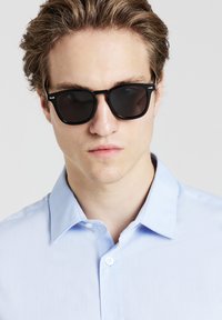 Occhiali da sole neri rettangolari con lenti scure, indossati con una camicia button-up azzurro chiaro e strutturata. La camicia presenta un colletto e un bottone singolo.