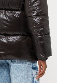 Μαύρο μπουφάν puffer με γυαλιστερή, πλισέ υφή, ελαστικούς καρπούς και χαλαρή γραμμή. Συνδυάζεται με ανοιχτό μπλε τζιν παντελόνι.