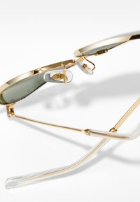 Gafas de sol redondas con montura de metal dorado y lentes verdes. Almohadillas nasales transparentes y puntas de las patillas, con un logotipo grabado sutil.