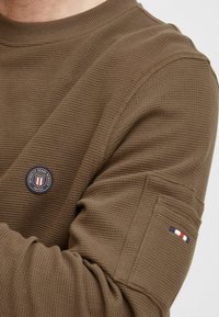Maglione marrone con texture, a girocollo, con un logo circolare sul petto e un'accorta rigata sulla manica.
