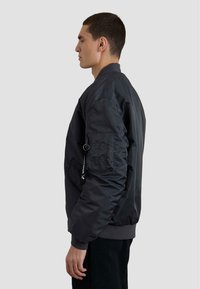 Giubbotto bomber nero con una consistenza liscia, polsini a coste e una tasca con zip sulla manica. Il design presenta una vestibilità comoda.