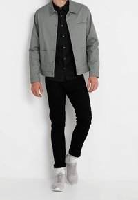 Grijze, lichtgewicht jas met drukknopen, gedragen over een zwart overhemd met knopen. Zwarte broek en grijze sneakers maken de outfit compleet.