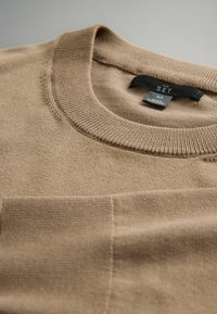 Beige Pullover aus weichem Stoff. Verfügt über einen gerippten Rundhalsausschnitt und eine Brusttasche, mit sichtbaren Steppdetails.
