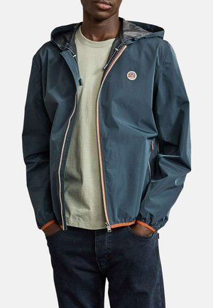 Homme portant une veste à capuche bleu marine zippée par-dessus un t-shirt vert clair et un jean foncé, les mains dans les poches de la veste.