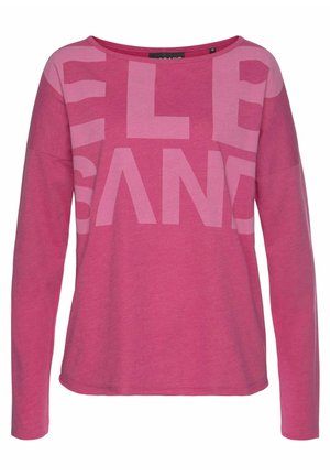 Chemise rose à manches longues avec un large décolleté et de grandes lettres en blocs rose clair partiellement visibles sur la partie supérieure de la poitrine et les manches.