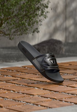 Melns slidens sandāls ar gludu, matētu virsmu. Aprīkots ar spīdīgu, abstraktu siksnas dizainu. Teksturēts zoles raksts pret koka dēļiem.