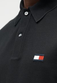 Μαύρο πόλο μπλουζάκι από υφασμάτινη υφή με πατιλέτα με τρία κουμπιά και μικρό κεντημένο λογότυπο με κόκκινες, λευκές και μπλε λεπτομέρειες στο στήθος.