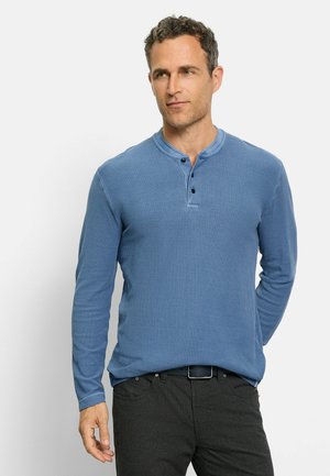 Langärmliges blaues Henley-Shirt aus strukturiertem Stoff mit Rundhalskragen, Knopfleiste und schwarzen Akzenten. Kombiniert mit dunklen Hosen.