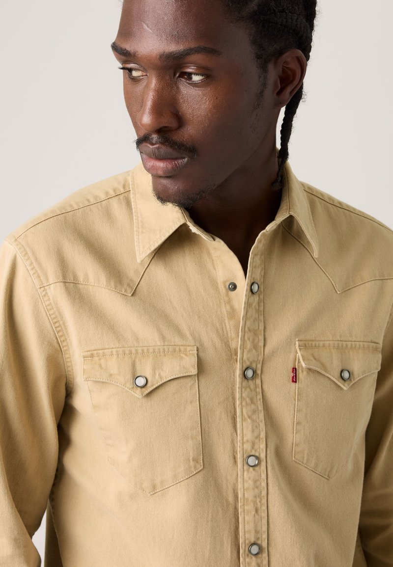 Homme aux cheveux tressés portant une chemise beige à boutons avec deux poches sur la poitrine, regardant vers sa gauche sur un fond neutre.