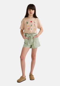 T-shirt brodé beige clair avec des motifs floraux, assorti à un short vert avec taille à nouer et poches ; sabots beiges aux pieds.