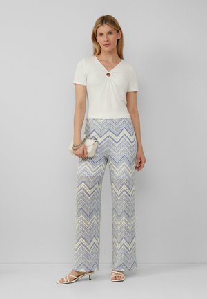 White fitted top with a center cutout, paired with wide-leg pants featuring a multicolored zigzag pattern in pastel tones. White clutch and sandals.
