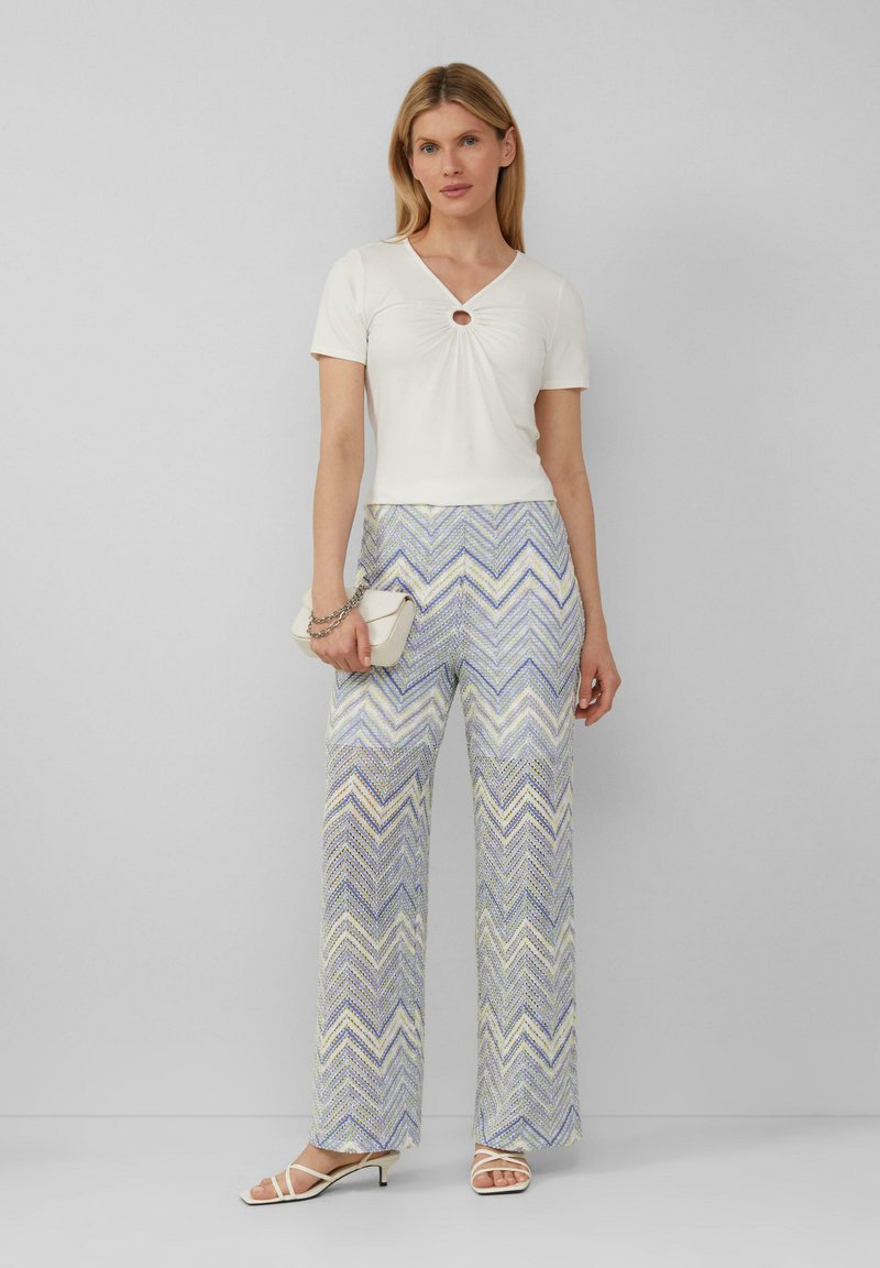 Witte fitted top met een middenuitsnijding, gecombineerd met een brede broek met een multikleurige zigzagpatroon in pasteltinten. Witte clutch en sandalen.