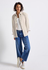 Vrouw in een beige corduroy overhemd met knopen over een witte top, blauwe wijde jeans en witte sneakers, staand tegen een effen achtergrond.
