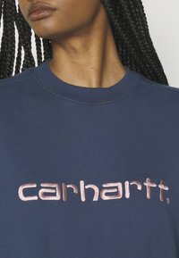 Sudadera navy con el logo "carhartt" bordado en rosa dorado, con cuello redondo acanalado y tejido texturizado.