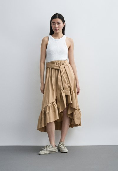 Γυναίκα που φοράει λευκό αμάνικο τοπ, μαύρη ασύμμετρη φούστα με βολάν και ζώνη, και μπεζ αθλητικά παπούτσια, στέκεται μπροστά σε γκρι απλό τοίχο.