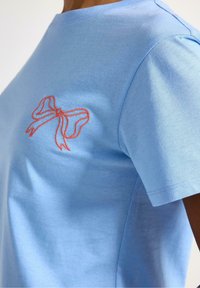 Vaaleansininen T-paita pehmeästä puuvillasta, jossa on oranssi brodeerattu rusetti vasemmassa rinnassa. Pyöreä kaula-aukko ja lyhyet hihat.