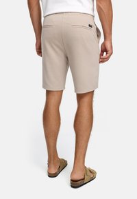 Hombre con pantalones cortos beige y sandalias marrones, de pie de espaldas contra un fondo blanco liso.