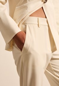Cremefarbenes, maßgeschneidertes Anzugensemble mit einem Blazer mit einem Knopf und gepressten Hosen; glatte Textur und strukturiertes Design.