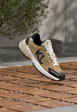 Beige and black Karl Lagerfeld sneaker with white laces and cushioning sole, suspended above terracotta tiles outdoors with greenery background.