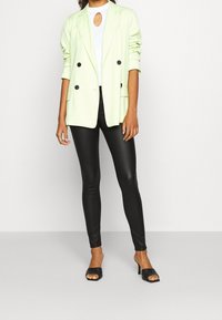 Blazer doble faz de color verde claro con botones negros, combinado con leggings negros. El modelo lleva una blusa blanca con escote de llave y mule negros con tacón.
