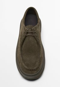 Chaussures en daim vert olive, dotées d'un bout arrondi, de coutures minimales et d'un système de laçage. La semelle est sombre et légèrement surélevée.