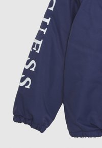 Námořnická puffy bunda s elastickým manžetovým rukávem, na boku s bílým nápisem "GUESS". Měkký, texturovaný materiál se lesklým povrchem.