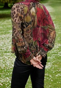 Blouse fluide et fleurie aux couleurs rouge, verte et noire ; manches longues avec poignets froncés ; tissu transparent ; accents en imprimé animal ; coupe loose.