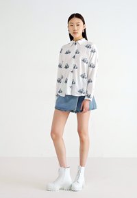White button-up shirt with black floral pattern, paired with a denim skirt and white platform ankle boots. Smooth fabric, relaxed fit.