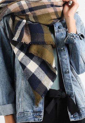 Écharpe à carreaux bleu marine, beige et olive drapée sur une veste en denim aux manches retroussées, portée avec un pantalon noir à taille nouée.