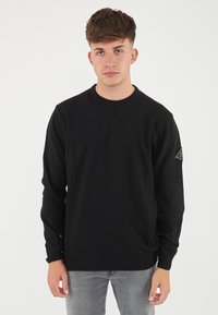 Maglione nero a collo rotondo realizzato in un morbido tessuto testurizzato, con maniche lunghe e un piccolo logo a forma di triangolo applicato sulla manica sinistra.