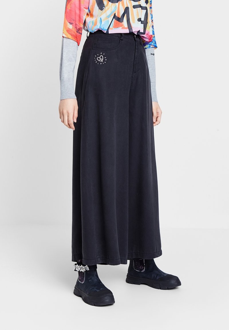 Personne portant une longue jupe noire avec un petit cœur blanc près de la poche, un haut coloré à manches longues et des bottines noires avec des détails floraux.