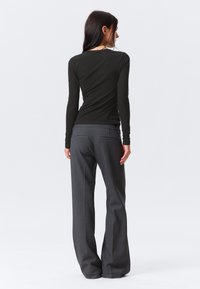 Kobieta stojąca tyłem, ubrana w czarną bluzkę z długim rękawem i szare szerokie spodnie, na jednolitym białym tle.