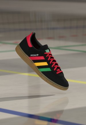 Baskets Adidas Spezial noires avec des lacets rouges, un talon rouge et des bandes jaunes, vertes et rouges, planant au-dessus du sol d'un terrain de sport avec des lignes colorées.